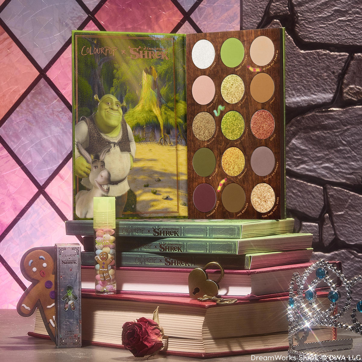 Shrek-Spress Yourself ColourPop Lip and Eye Set showcasing an open 15-pan green and neutral eyeshadow palette with Shrek artwork and a gingerbread-man lip gloss, posed on stacked storybooks by a castle window with tiara and rose.