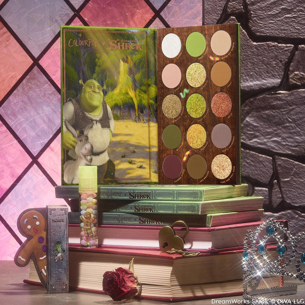 Shrek-Spress Yourself ColourPop Lip and Eye Set showcasing an open 15-pan green and neutral eyeshadow palette with Shrek artwork and a gingerbread-man lip gloss, posed on stacked storybooks by a castle window with tiara and rose.