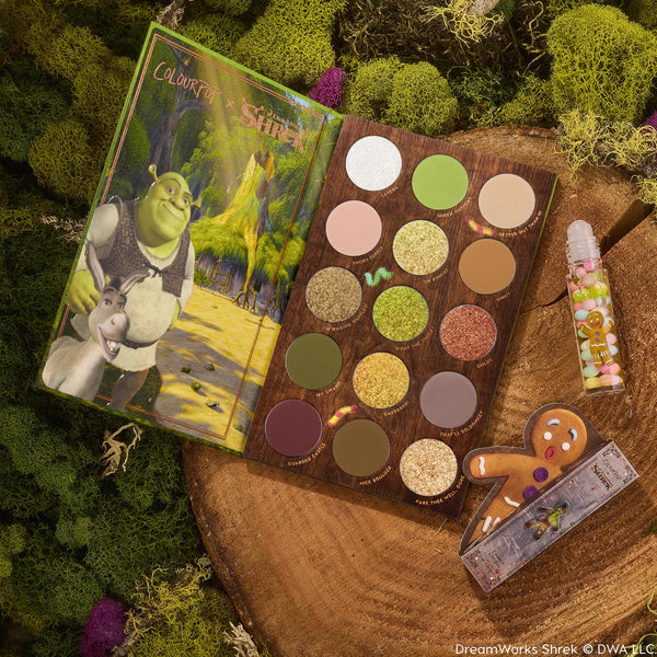 Shrek-Spress Yourself ColourPop Lip and Eye Set showcasing an open 15-pan eyeshadow palette of greens, golds, and glittery neutrals, plus a clear sprinkle lip gloss and gingerbread man accessory, styled on mossy wood for a swamp-inspired vibe.