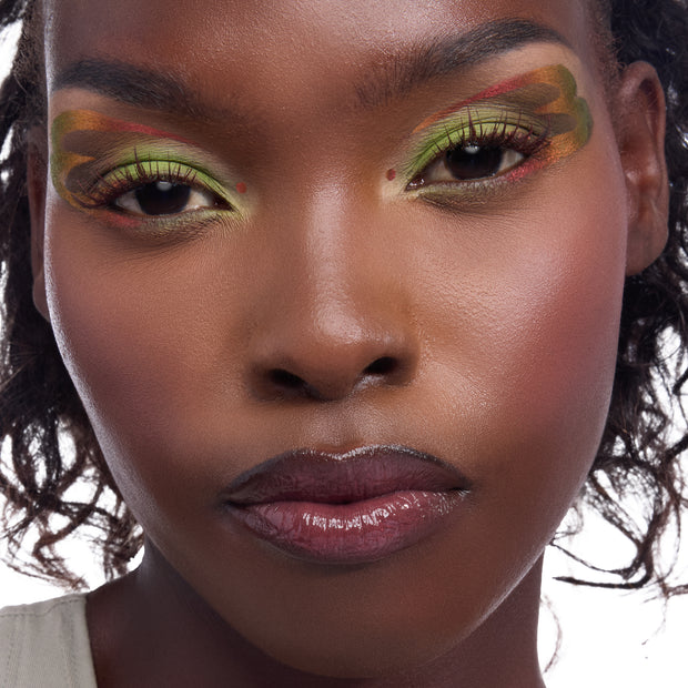 Dragon’s Keep ColourPop Crème Gel Liner creating bold lime‑green and rusty‑red graphic wings with inner‑corner dots on a close‑up model’s eyes, showcasing rich pigment and long‑wear detail against glowy skin in a studio shot.