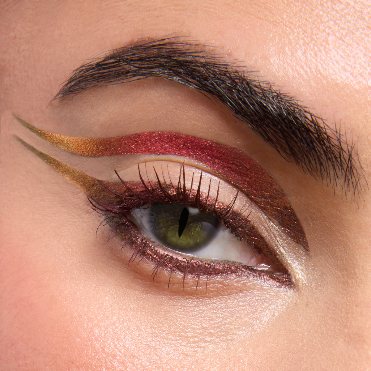 Eye showcasing Dragon’s Keep Crème Gel Liner by ColourPop Crème Gel Liner in a bold double-wing - metallic red-to-copper arc above the crease, olive-gold swoop extending outward, and shimmering rose along the lashline - on fair skin with a dark brow.
