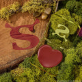 Fairytale Things ColourPop Lip and Cheek Balm sits open, revealing a heart-shaped pan of warm brick-red pigment in a translucent green, Shrek-inspired compact with an ogre-ear S, resting on moss and a wood slice beside a glossy burgundy S swatch.