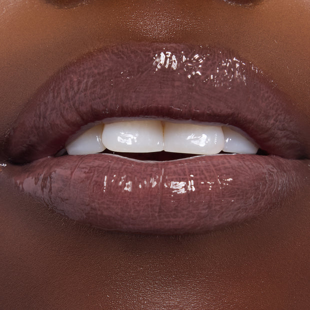 Glossy lips wearing ColourPop Ultra Glossy Lip in Noble Steed, a deep mauve-brown shade, shining with a high-shine finish and smooth, even coverage; close-up beauty swatch on dark skin with teeth slightly parted.