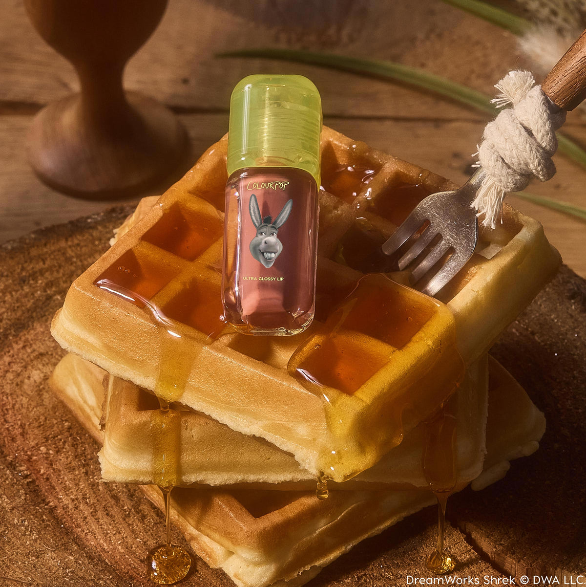 ColourPop Ultra Glossy Lip in Noble Steed rests on syrup-drizzled waffles, the compact gloss bottle featuring a lime-green cap and Donkey graphic label, styled on a rustic wooden board with a fork for a playful breakfast-themed product shot.