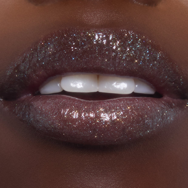 Lips wearing ColourPop Ultra Glossy Lip in Ogre, catching light with multi-dimensional shimmer and glassy shine as teeth gently show; studio macro highlights the deep plum base packed with fine glitter.