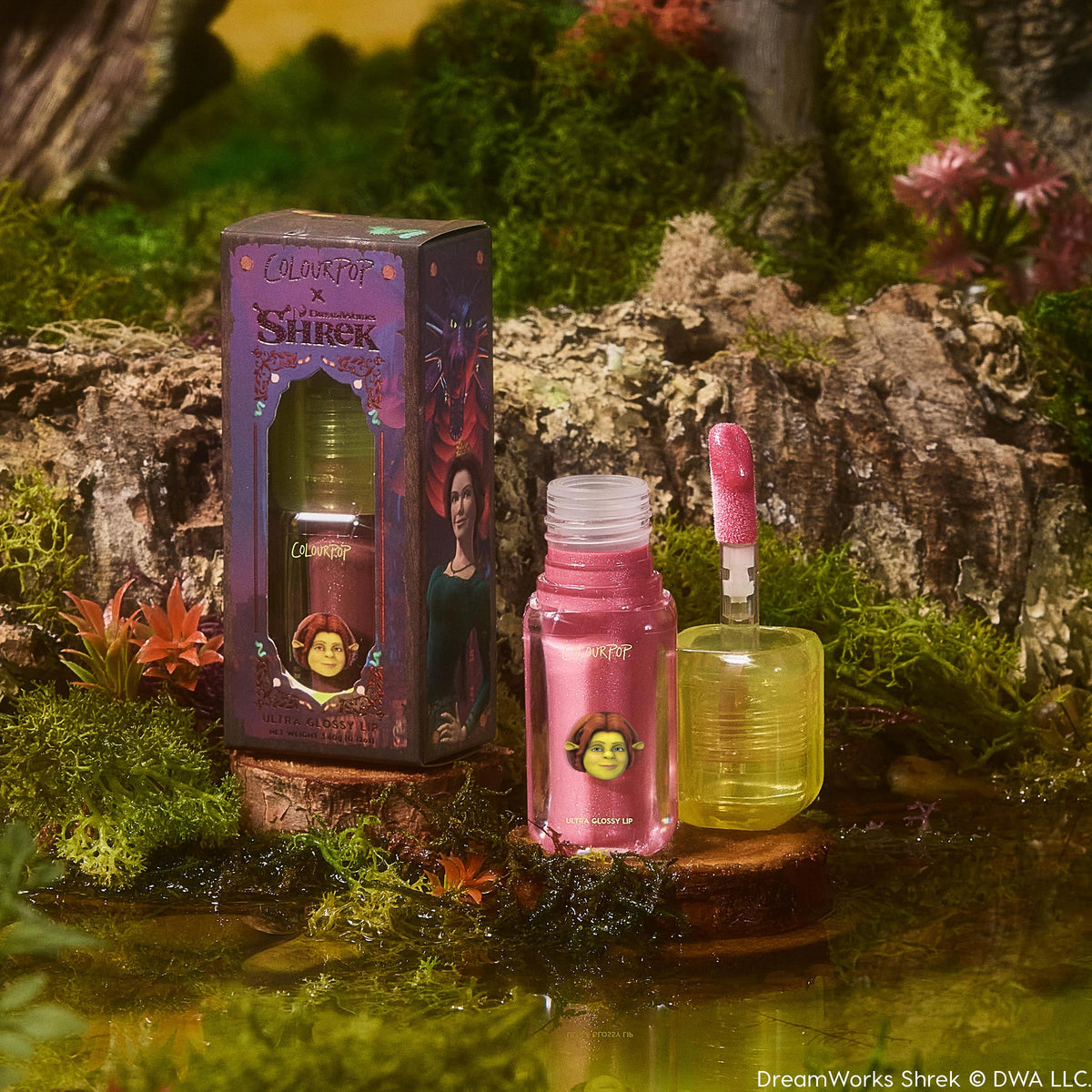 ColourPop Ultra Glossy Lip in Ogress shown with an open sparkly pink gloss tube and doe-foot applicator beside the Shrek x ColourPop illustrated box, styled on mossy swamp scenery for a whimsical look.