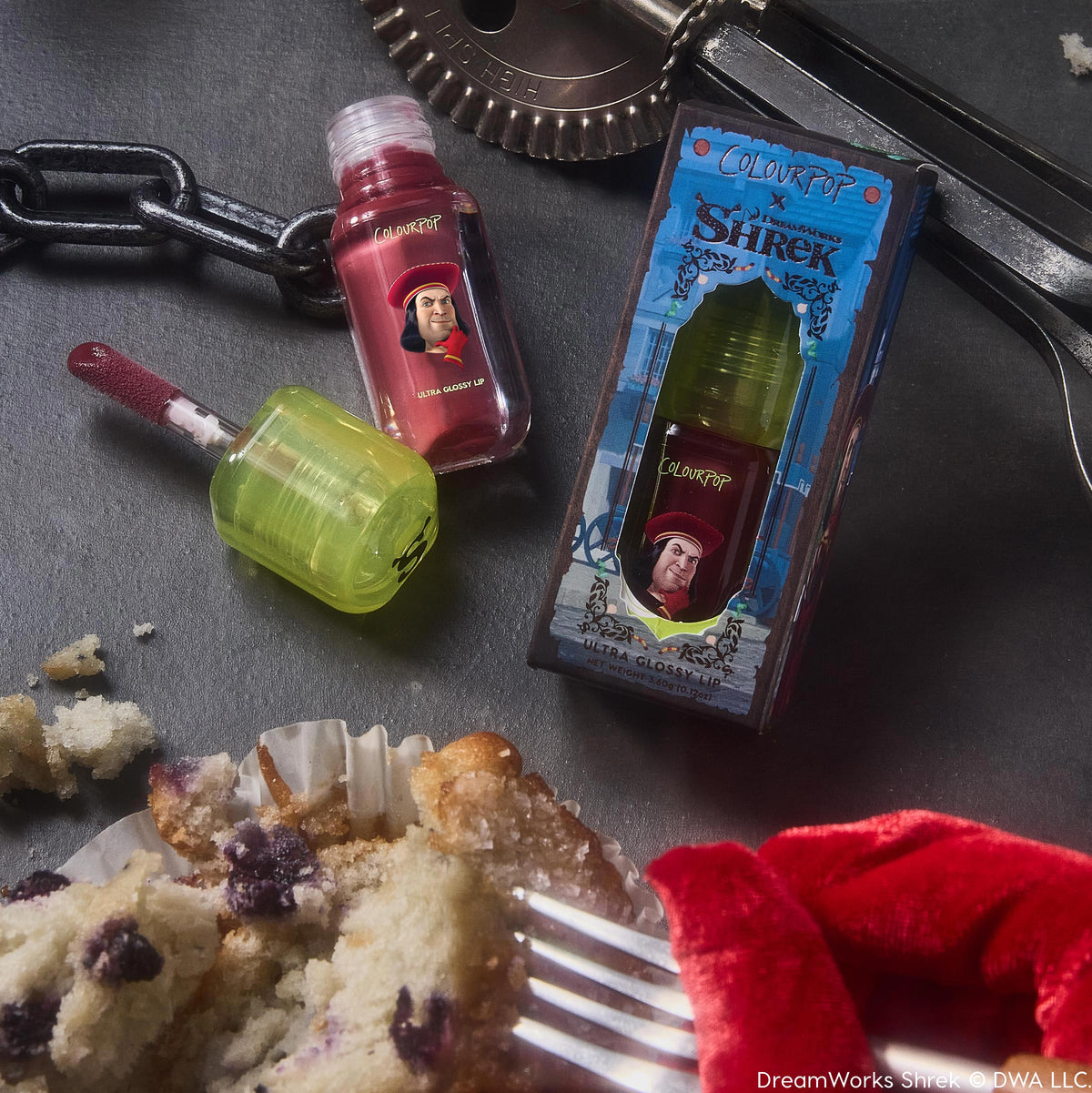 Short Lord ColourPop Ultra Glossy Lip - red tint with doe-foot applicator and neon green cap - rests open beside its Shrek-themed box featuring Lord Farquaad on a dark tabletop with crumbs, chain, and kitchen tools.