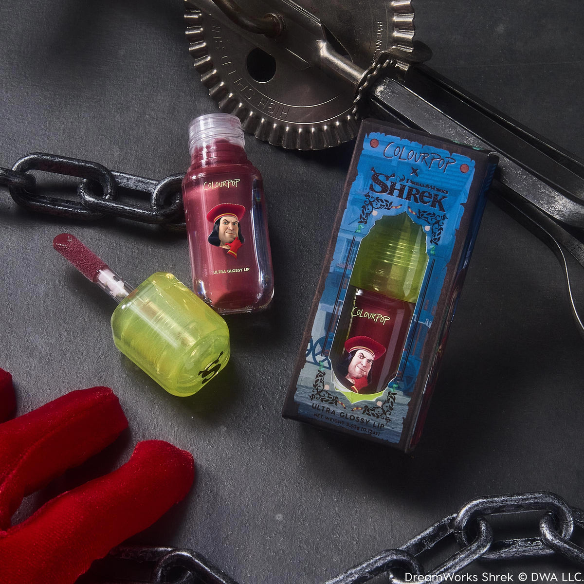 ColourPop Ultra Glossy Lip “Short Lord” with lime-green cap and open doe-foot applicator shows a deep berry gloss and Lord Farquaad art beside the Shrek box, set on a dark stone surface with chains, metal gears, and a red glove.