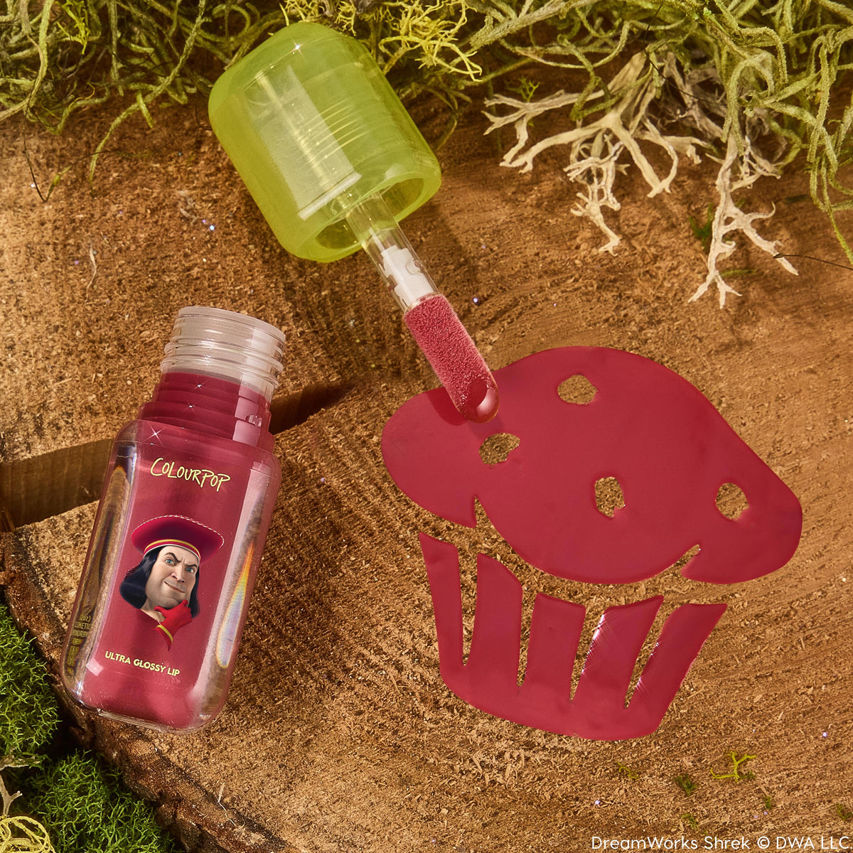 Short Lord Ultra Glossy Lip by ColourPop, applicator swatching raspberry-pink gloss through a cupcake stencil on a wood stump with moss; translucent green cap and bottle art featuring Lord Farquaad from Shrek; ColourPop Ultra Glossy Lip.
