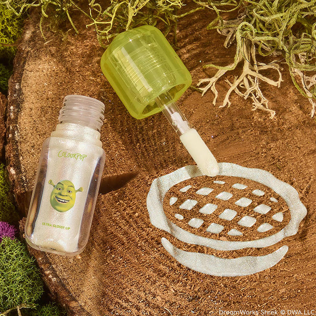 ColourPop Ultra Glossy Lip ‘Ogre’ shown open, doe‑foot applicator swiping pearlescent shimmer gloss in a swatch on a wood slice; clear tube with Shrek graphic and lime green cap, surrounded by moss for a swamp‑inspired product shot.