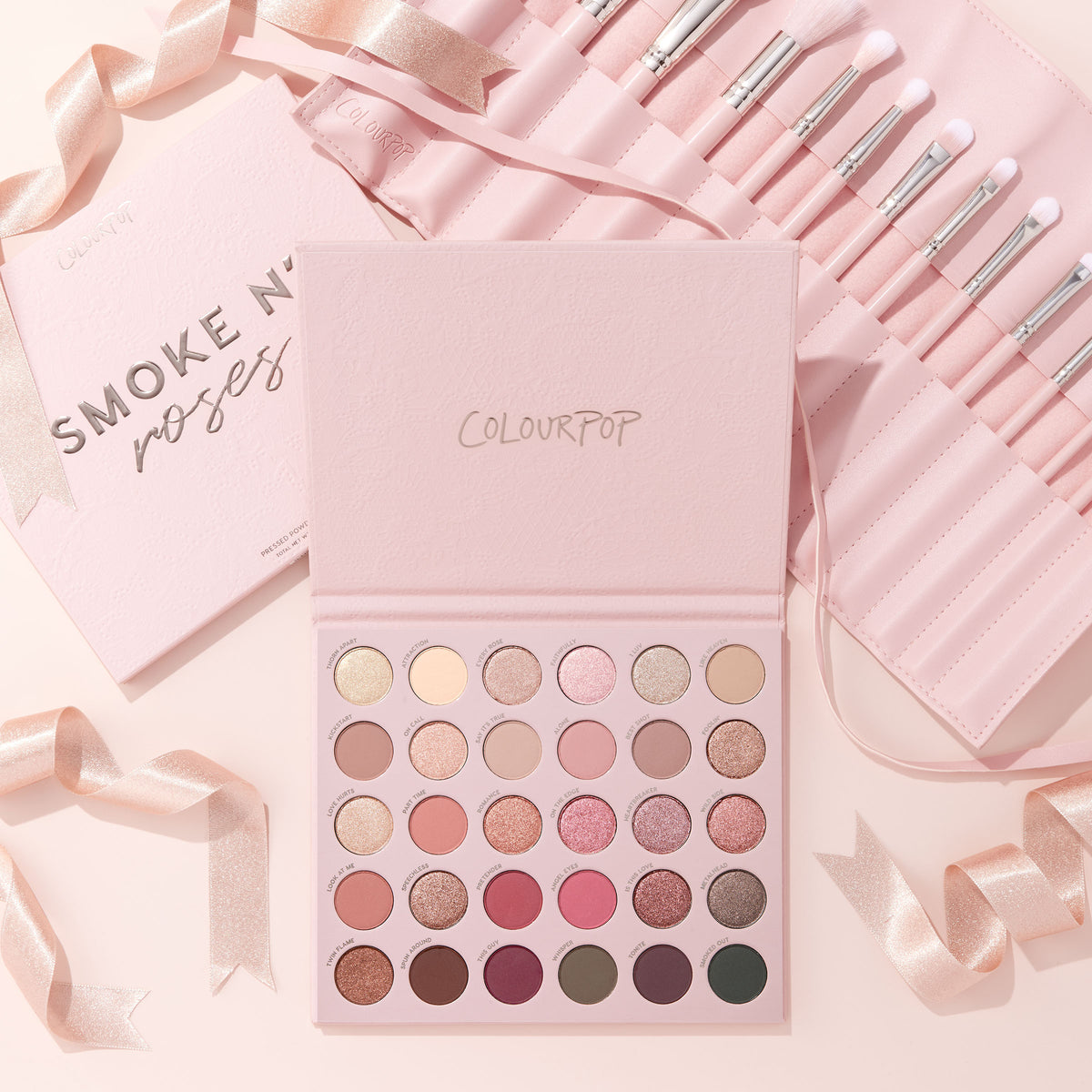 Total Smokeshow ColourPop Shadow Palette & Brush Set displaying rows of rosy neutrals and shimmering tones beside a pink roll of white‑tipped brushes, styled on a soft blush backdrop with satin ribbons.