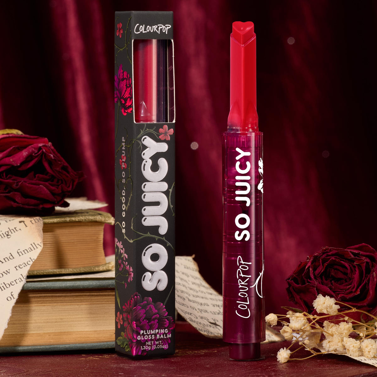 ColourPop So Juicy Plumping Gloss in Spice Level stands upright with a heart‑tipped stick, showing a deep berry red shade beside its black floral box, styled with vintage books and dried roses against a burgundy velvet backdrop.