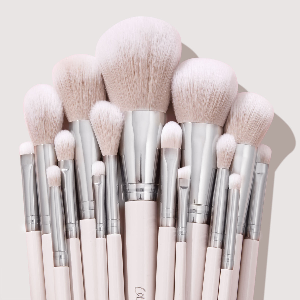 Soft pink makeup brushes with silver ferrules fan upright, showcasing powder, blush, and eyeshadow heads in a neat cluster on a neutral backdrop, part of The Ultimate Stunner ColourPop Shadow Palette & Brush Set for precise application.