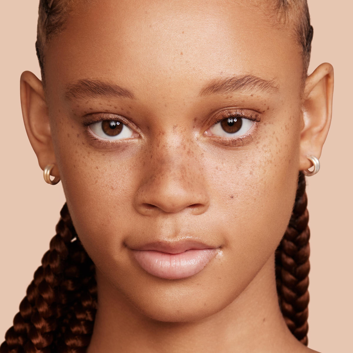 Model wears Medium 100 W shade of ColourPop Hyaluronic Hydrating Foundation, showcasing even, dewy, skin-like coverage that lets freckles show through; close-up face with braided hair and hoop earrings on a beige studio background.