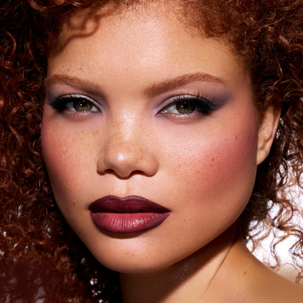 Model wearing ColourPop New Love Lippie Stix from the Twilight collection, showing rich matte deep berry lipstick on full lips with soft-focus glam eye makeup in a close-up beauty shot against a clean studio background.