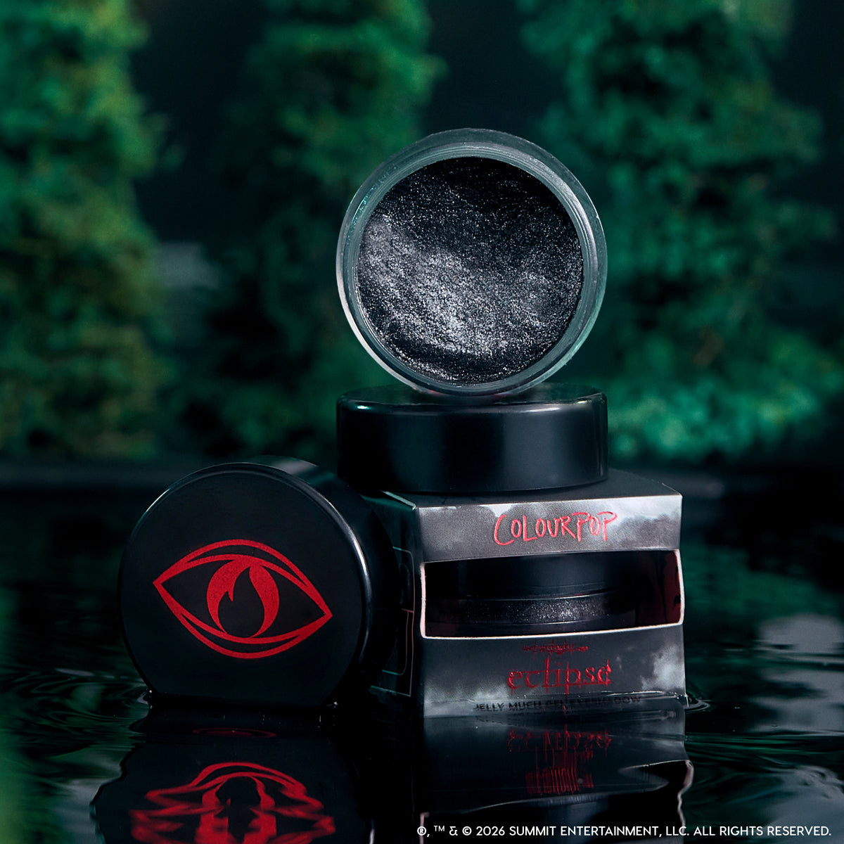 Open pot of ColourPop Newborn Army Jelly Much Shadow showing deep black, silver-flecked Twilight-inspired gel eyeshadow stacked with eclipse collection packaging and red eye logo, reflected on dark water against blurred forest background.