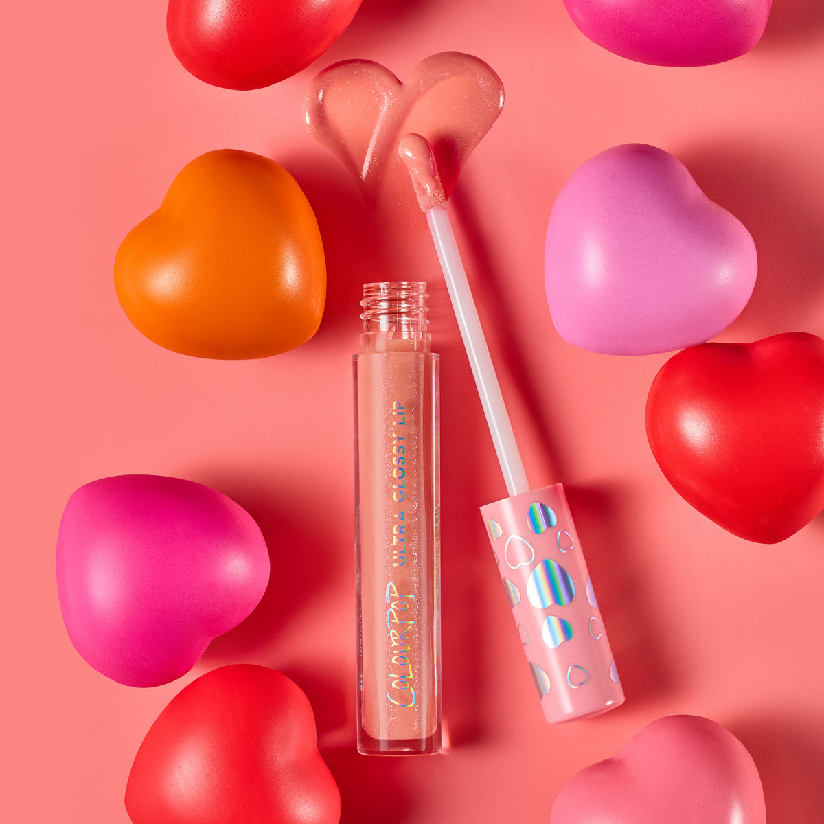 ColourPop Ultra Glossy Lip in One Kiss, a peachy‑pink shimmer, shown with open tube and applicator creating a heart-shaped swipe; pink cap with iridescent hearts; surrounded by red, pink, and orange heart props on a coral background.