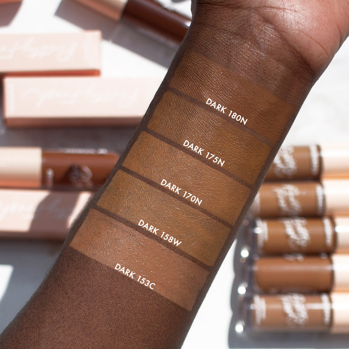 ColourPop Pretty Fresh Creamy Concealer in Dark 153 C swatched on a forearm as the bottom labeled block, shown with neighboring deep shades (158W, 170N, 175N, 180N); concealer tubes and pink boxes sit in bright sunlight.
