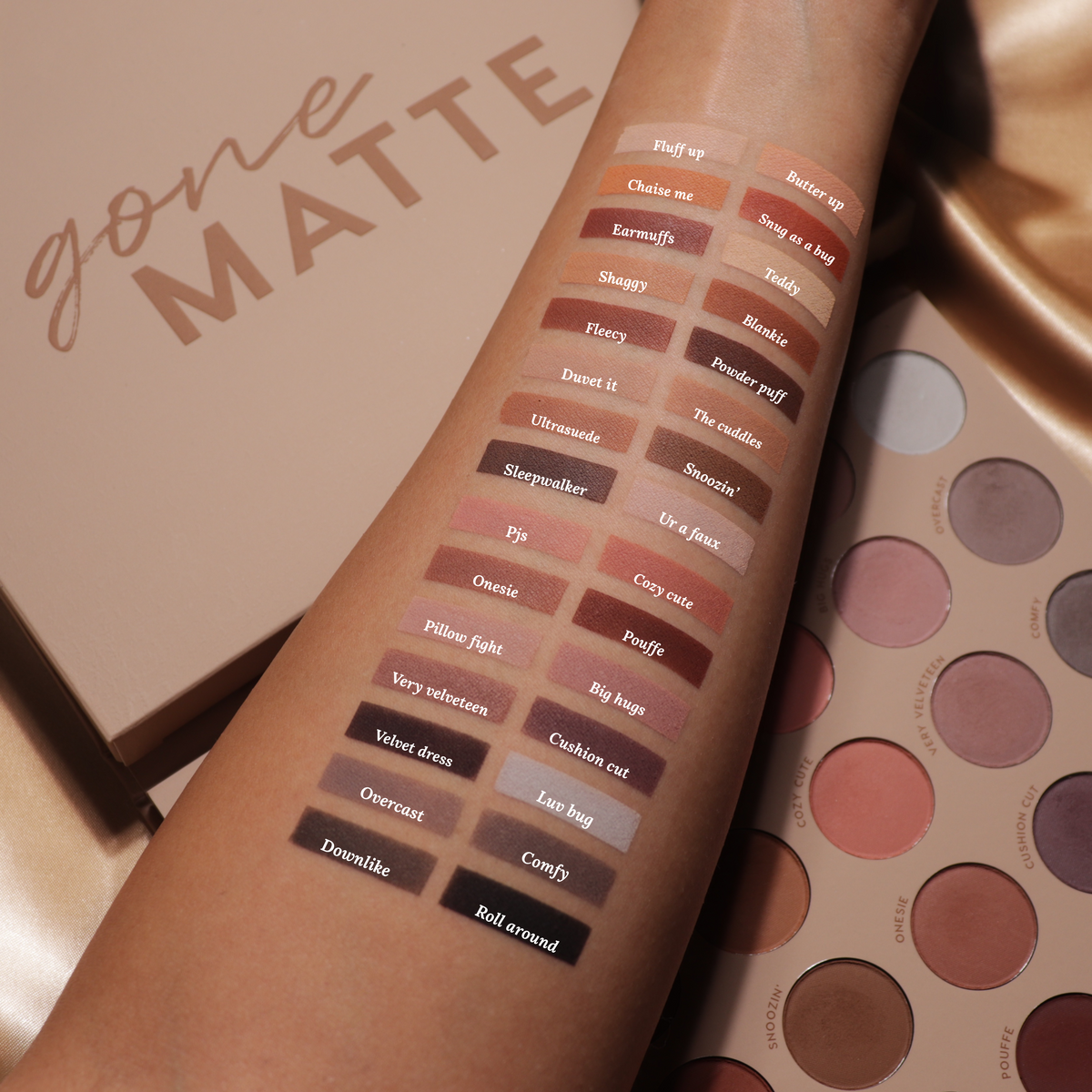 ColourPop Gone Matte shadow palette swatches shown on a forearm, each shade name printed beside matte nudes, taupes, pinks, and deep browns to black, while the open palette and matching tan box sit nearby on a warm studio backdrop.