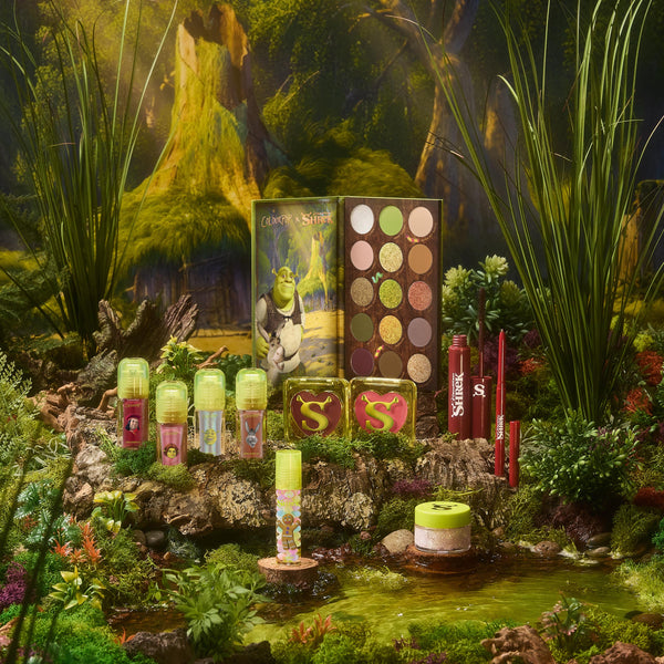 ColourPop Shrek collaboration cosmetics displayed on a mossy log, featuring a 15-pan green and neutral eyeshadow palette with Shrek art, mini lip glosses, two heart-shaped blush compacts, red lip products, and glitter in a whimsical swamp scene.