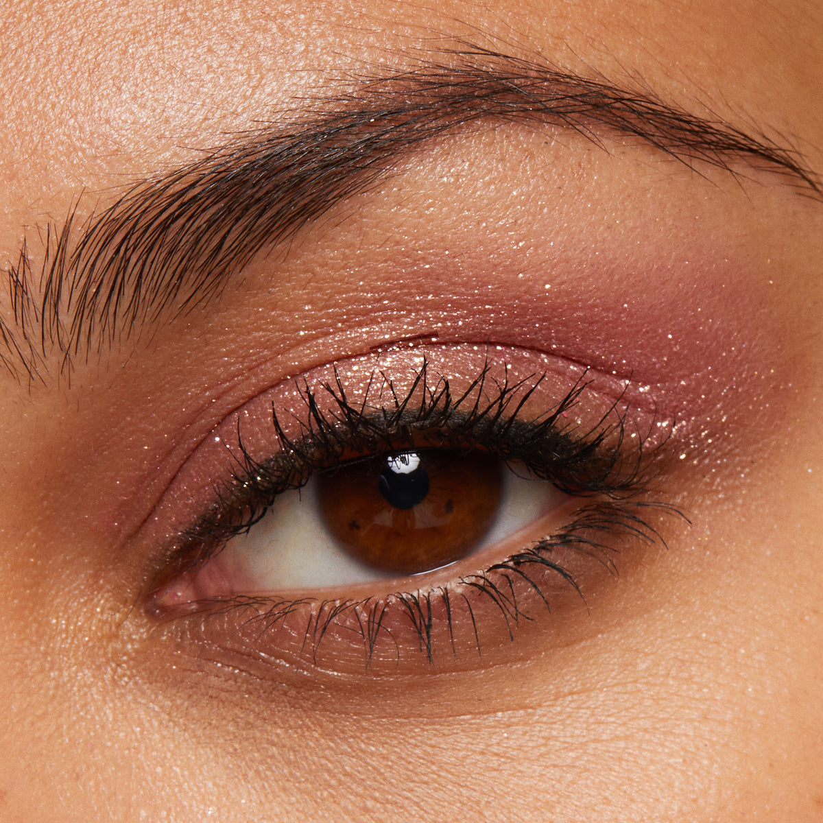 ColourPop Roy G. Biv Super Shock Shadow glitters on the eyelid, delivering a rosy pink base with gold sparkle and a creamy metallic finish, shown in a macro close-up of a brown eye with lashes and brow.