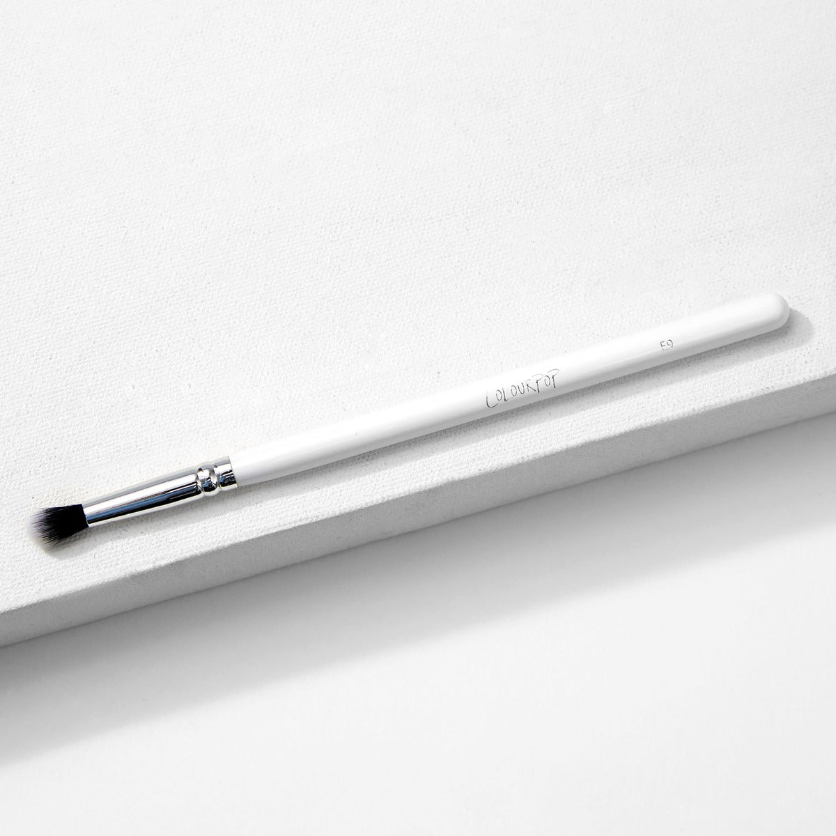 ColourPop Small Tapered Brush resting diagonally on a white textured platform, featuring a slim white handle, chrome ferrule, and small tapered black-and-white bristles for precise eyeshadow blending and detail work.