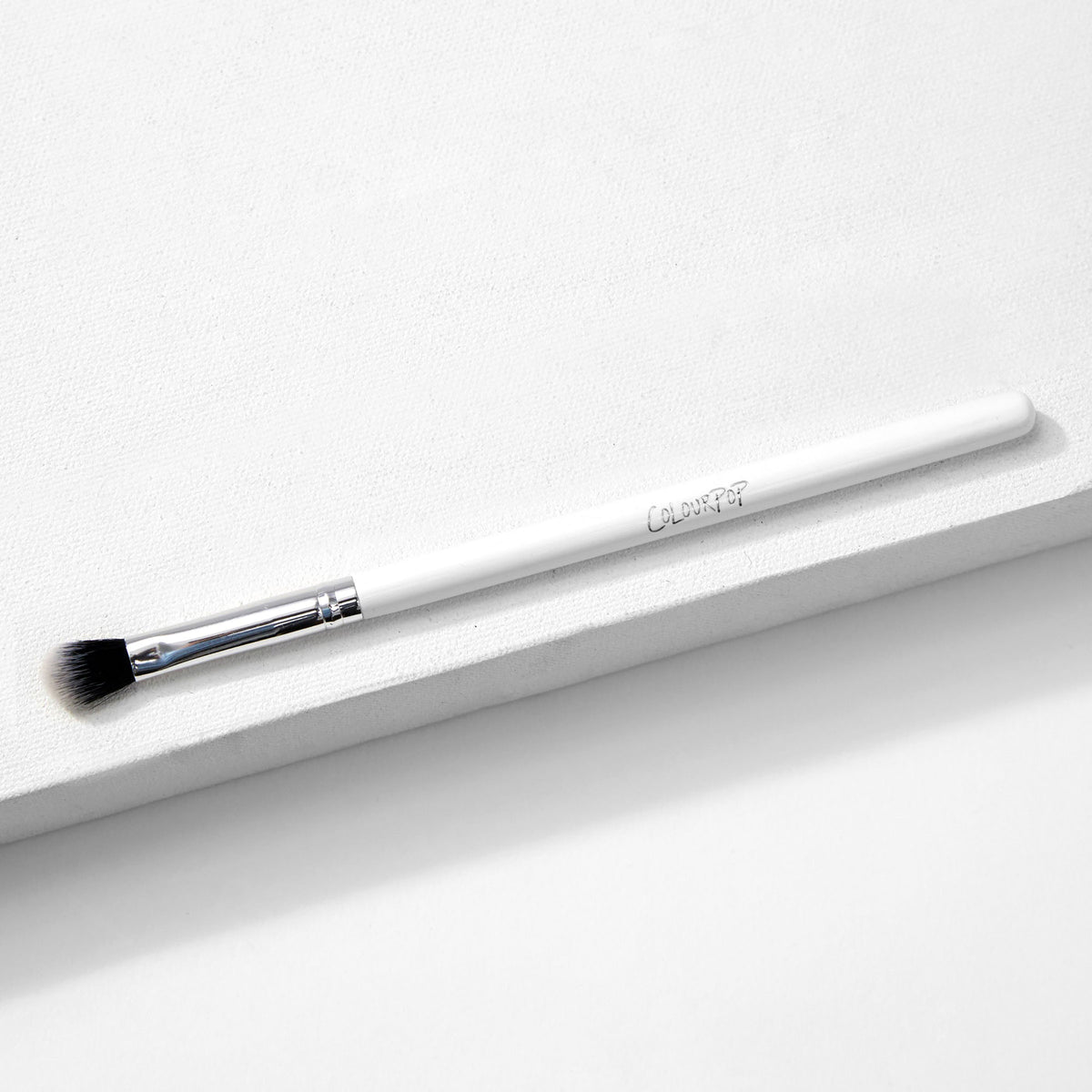 ColourPop Blending Brush makeup brush resting diagonally on a white textured surface, featuring fluffy angled black-to-white bristles, a silver ferrule, and a slim white handle, made for seamless eyeshadow blending.