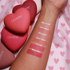 ColourPop Kiss 'N Tell pressed powder blush swatched on a forearm, with six labeled shades - Kiss N Tell, Let’s Dance, Flirt Alert, 4ever Yours, Text Me, Babycakes - beside matte heart-shaped ColourPop compacts on pink heart-print fabric.