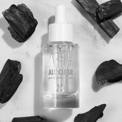 Glass dropper bottle of Fourth Ray Beauty All Clear Purifying Serum standing upright, showcasing clear formula and label, surrounded by charcoal pieces on a white marble surface; .85 fl oz/25 mL ColourPop Face Serum for purifying skin.