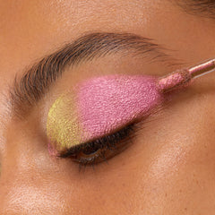 Doe-foot applicator sweeping ColourPop Chrome Liquid Shadow in Bliss across a closed eyelid, showing a metallic duochrome shift from lime-gold at the inner corner to vivid pink over the lid in a close-up beauty shot.