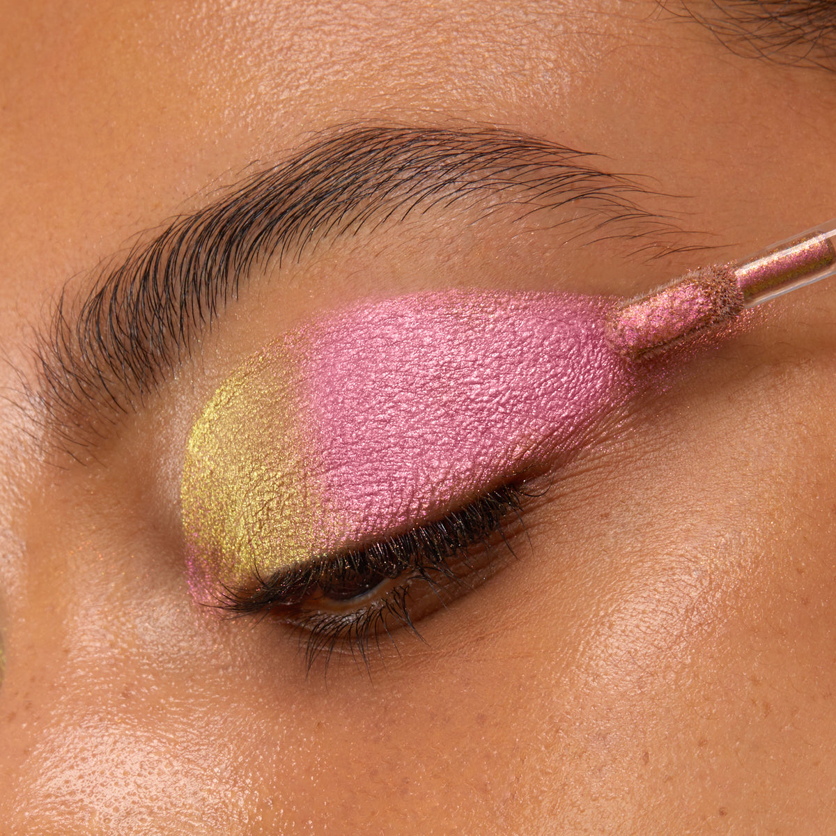 Doe-foot applicator sweeping ColourPop Chrome Liquid Shadow in Bliss across a closed eyelid, showing a metallic duochrome shift from lime-gold at the inner corner to vivid pink over the lid in a close-up beauty shot.