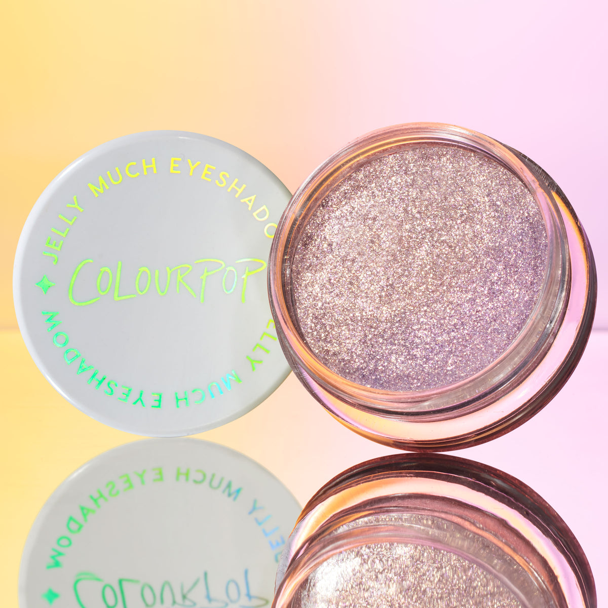 ColourPop Jelly Much Shadow Boo-Kay shown open, a sparkling lavender-lilac gel eyeshadow in a clear round pot, white lid with neon green logo beside it, reflected on a mirror with a pink-to-yellow gradient background.