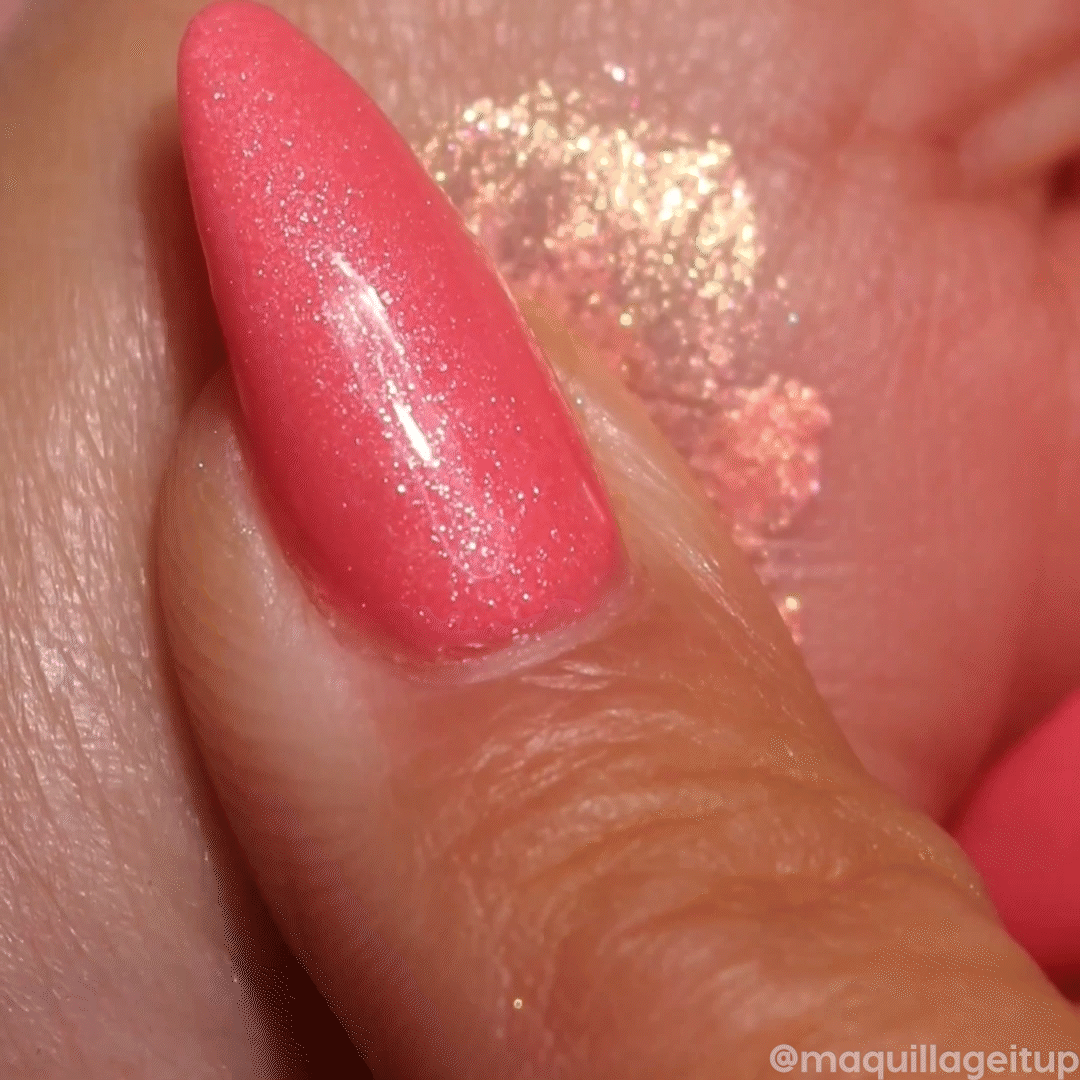 ColourPop Jelly Much Shadow Buzzy swatch - sparkling peach-gold gel eyeshadow with pink-coral shimmer - being swiped on skin by a coral-painted fingertip, showcasing a high-shine metallic finish and bold, long-wear payoff.