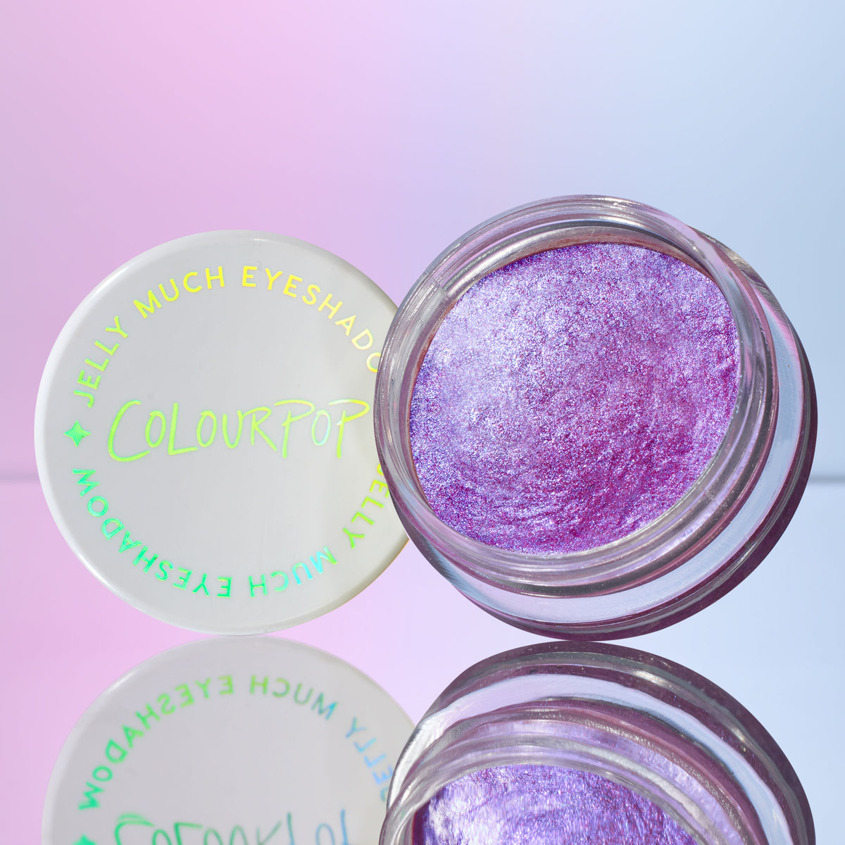 Open jar of ColourPop Jelly Much Shadow, Duckling, revealing metallic lavender‑pink shimmer in a clear pot, holographic ColourPop lid beside it, styled on a reflective surface with a pastel blue‑pink gradient backdrop.