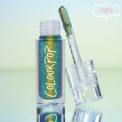 Beetle ColourPop Chrome Liquid Shadow stands open with a doe-foot applicator resting on the clear star-etched cap, revealing a multichrome green, teal, and gold shift in a clear tube on a reflective surface with a pastel gradient backdrop.