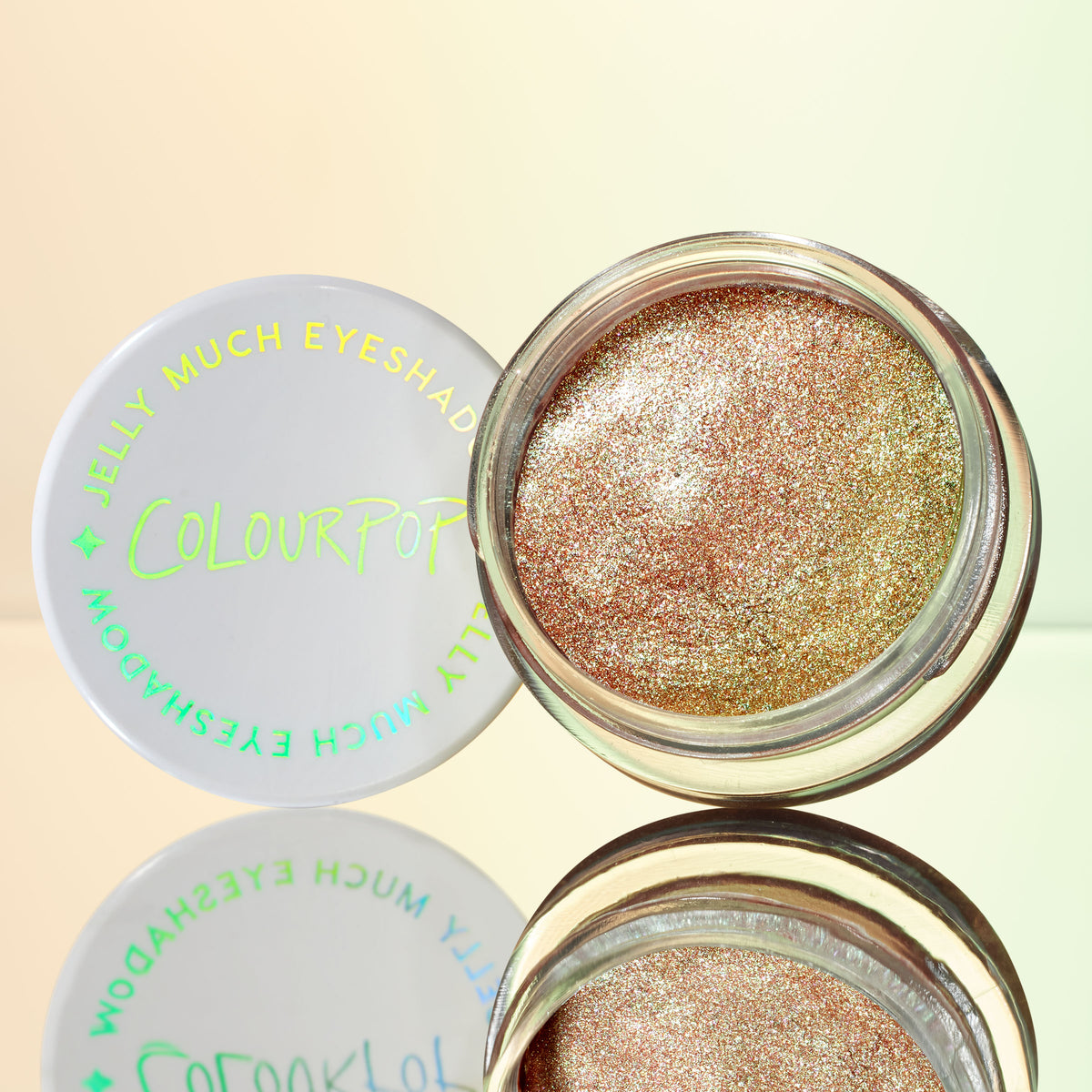 Open pot of ColourPop Jelly Much Shadow in Shiny Penny glimmers with metallic copper-rose shimmer, while its white lid with holographic logo rests beside it on a reflective surface against a soft gradient backdrop.