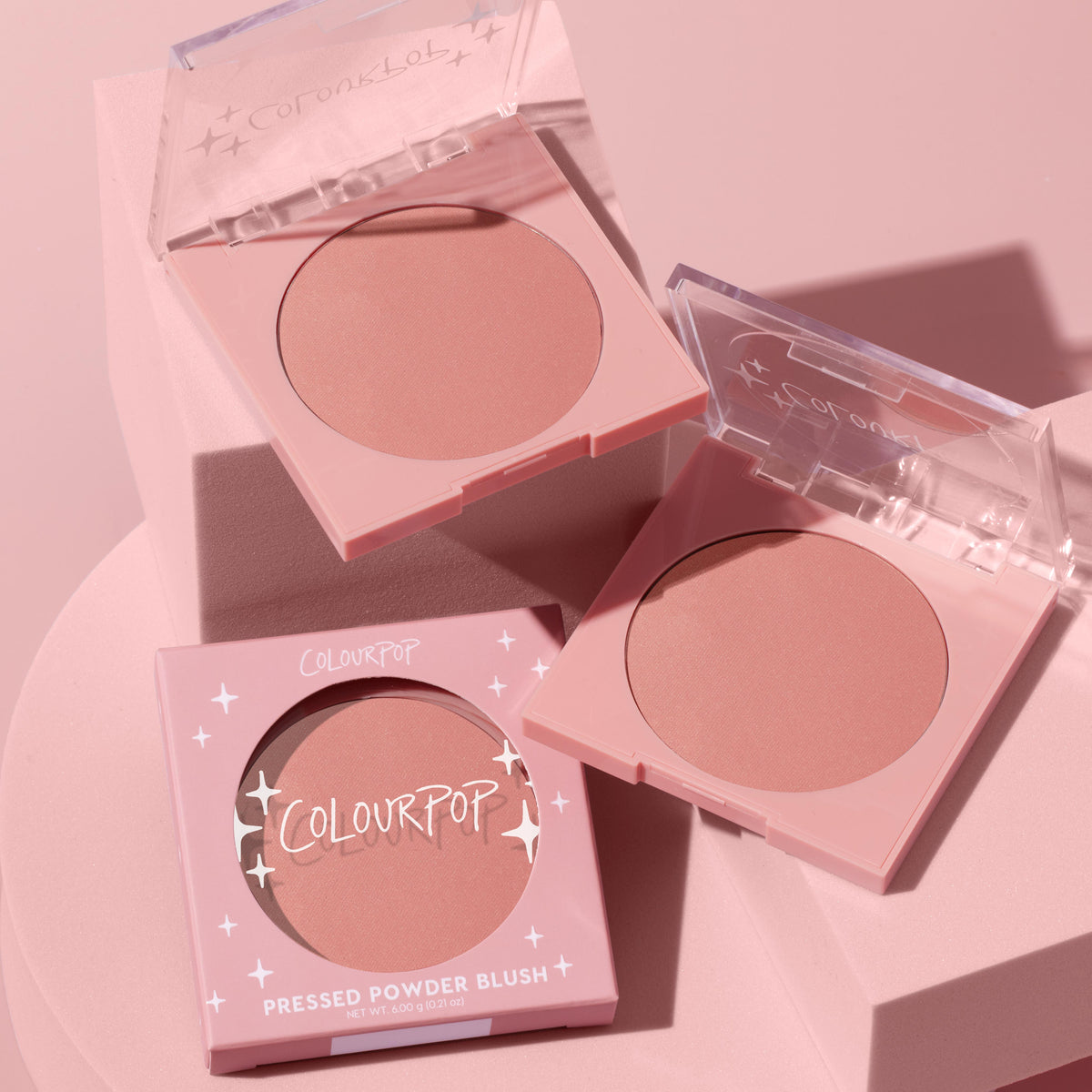 To the 10 ColourPop Pressed Powder Blush in pastel pink compacts, opened to show a soft rosy nude shade, styled with its boxed packaging on a pink pedestal under soft studio lighting, 6.0 g (0.21 oz) pan.