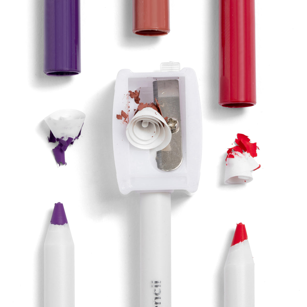 ColourPop Pencil Sharpener trimming creamy purple and red cosmetic pencils, curled shavings visible beside capped lip and eye crayons on a clean white background; a Makeup Tools & Accessories essential for sharp, precise tips.