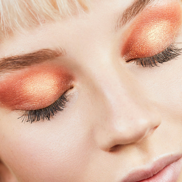ColourPop Super Shock Shadow in Pledge Class shimmering across closed eyelids, showing a warm copper-to-peach gradient with golden metallic sparkle and soft lashes in a close-up beauty shot.